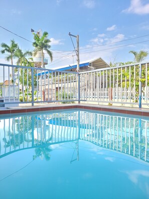Une piscine entourée de palmiers et bordée d’une rambarde en métal, avec un bâtiment aux toits bleus à proximité.