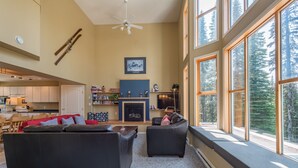 A spacious living room with a fireplace, large windows, and a view of the forest. - Amazing location right on the ski way - bright home with private hot tub (Silver Star Mountain)