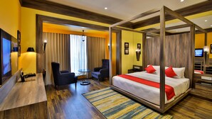 A hotel room with a four-poster bed, a desk with a chair, a TV, and a seating area with a table and chairs.