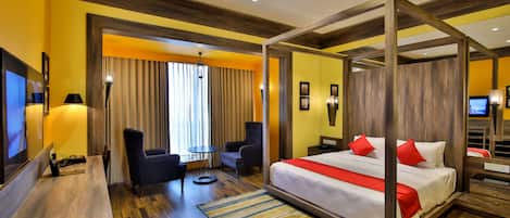 A hotel room with a four-poster bed, a desk with a chair, a TV, and a seating area with a table and chairs.