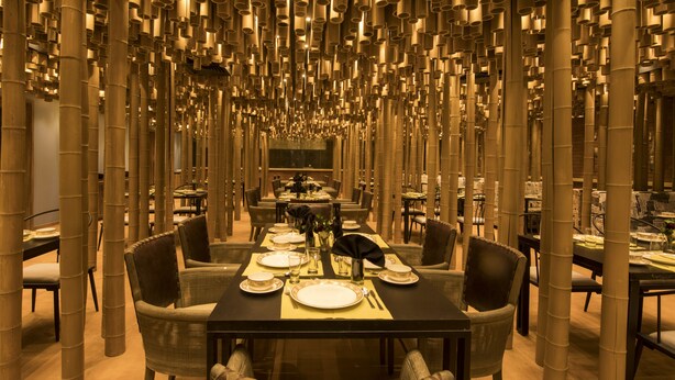 A restaurant with a long table set for dining, surrounded by chairs, and a ceiling with cylindrical structures. - Sayaji Rajkot (Rajkot)