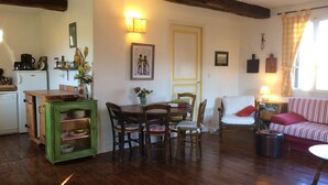 Una sala acogedora con suelo de madera, una mesa de comedor con sillas, un mueble verde, un sofá y una mesita con una lámpara.