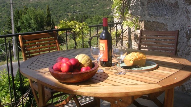 Una mesa de madera con un bol de manzanas rojas, una botella de vino y una hogaza de pan en un balcón con vistas a un paisaje montañoso.