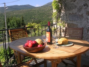 Een houten tafel met een schaal rode appels, een fles wijn en een brood op een balkon met uitzicht op een bergachtig landschap.