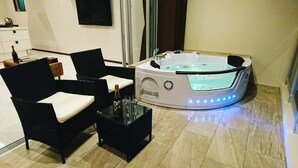 Un baño moderno con hidromasaje, sillas negras, una mesa de cristal con una botella y vasos, y un televisor de pantalla plana en la pared.