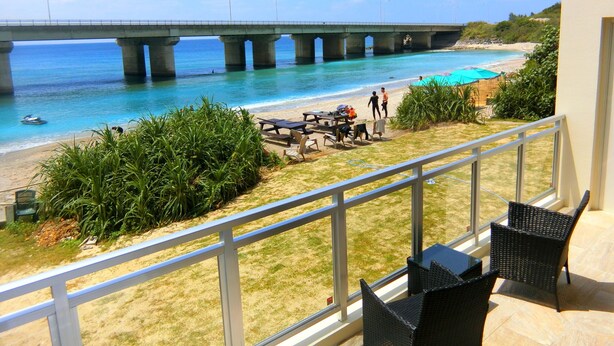 Un balcón con sillones de mimbre que da a una playa, con un puente al fondo.