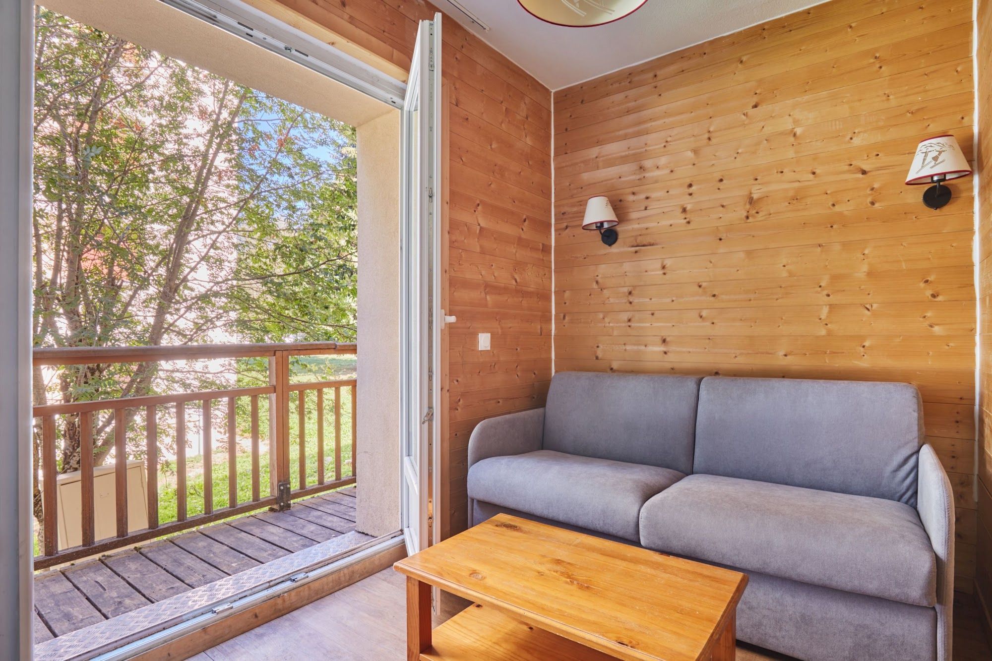 A wooden interior with a grey sofa and a wooden table, a balcony with a wooden railing, and two wall-mounted lamps.