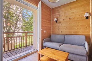 A wooden interior with a grey sofa and a wooden table, a balcony with a wooden railing, and two wall-mounted lamps.
