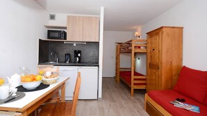 Quarto compacto com cozinha compacta, uma mesa de jantar com itens de café da manhã, uma cama beliche com roupa de cama vermelha e um armário de madeira.