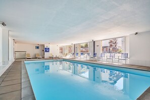 Una piscina interior con paredes de vidrio transparente, suelo de baldosas y varias sillas de descanso.