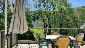 Een balkon met een tafel en stoelen, een hangende parasol en uitzicht op bomen en een meer.
