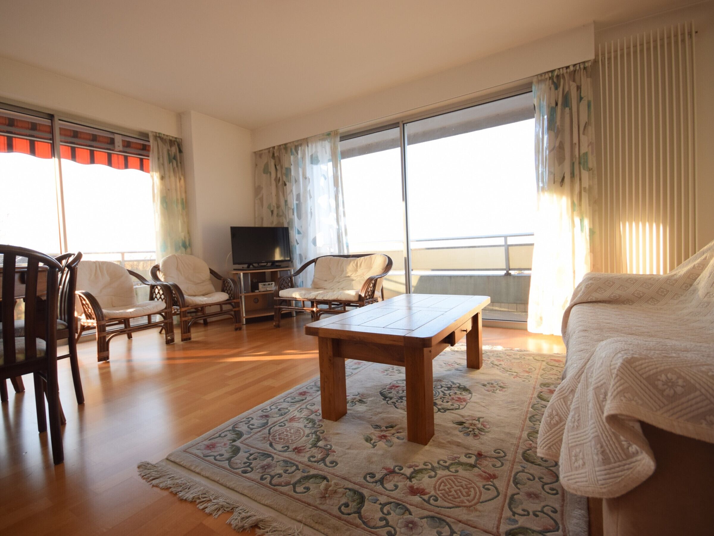 Un salón con una mesa de centro de madera, dos sillones, un sofá, un televisor sobre un mueble bajo y un ventanal con cortinas.