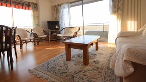 Un salon avec une table basse en bois, deux fauteuils, un canapé, un téléviseur posé sur un meuble et une grande fenêtre avec des rideaux.