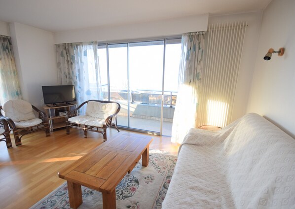 A living room with a wooden coffee table, two wicker chairs, a sofa, a television, and a large sliding glass door leading to a balcony.
