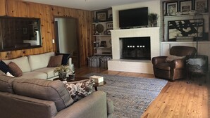 A cozy living room with a fireplace, wooden walls, a sofa, a coffee table, and a TV mounted above the fireplace. - Quiet Home less than a mile from Vaught Hemingway (Oxford)