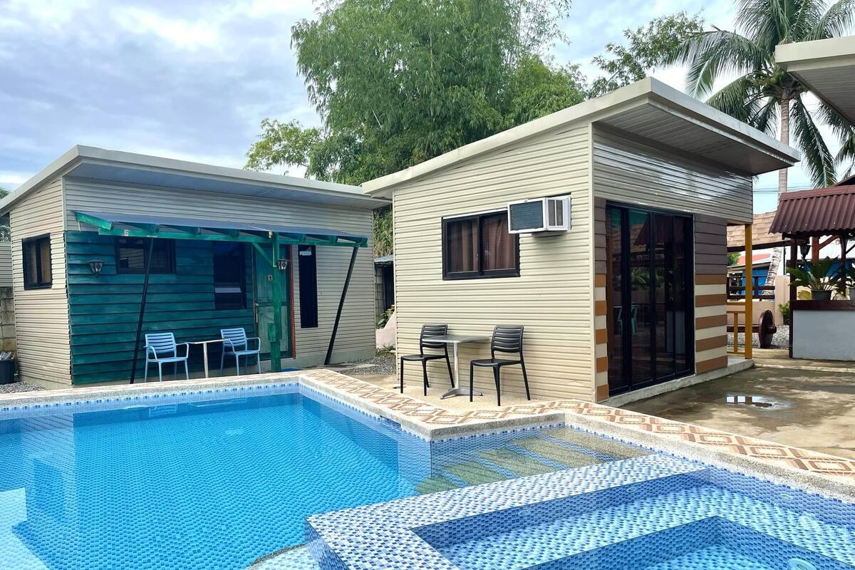 A pool area with a rectangular pool and a covered seating area with chairs.