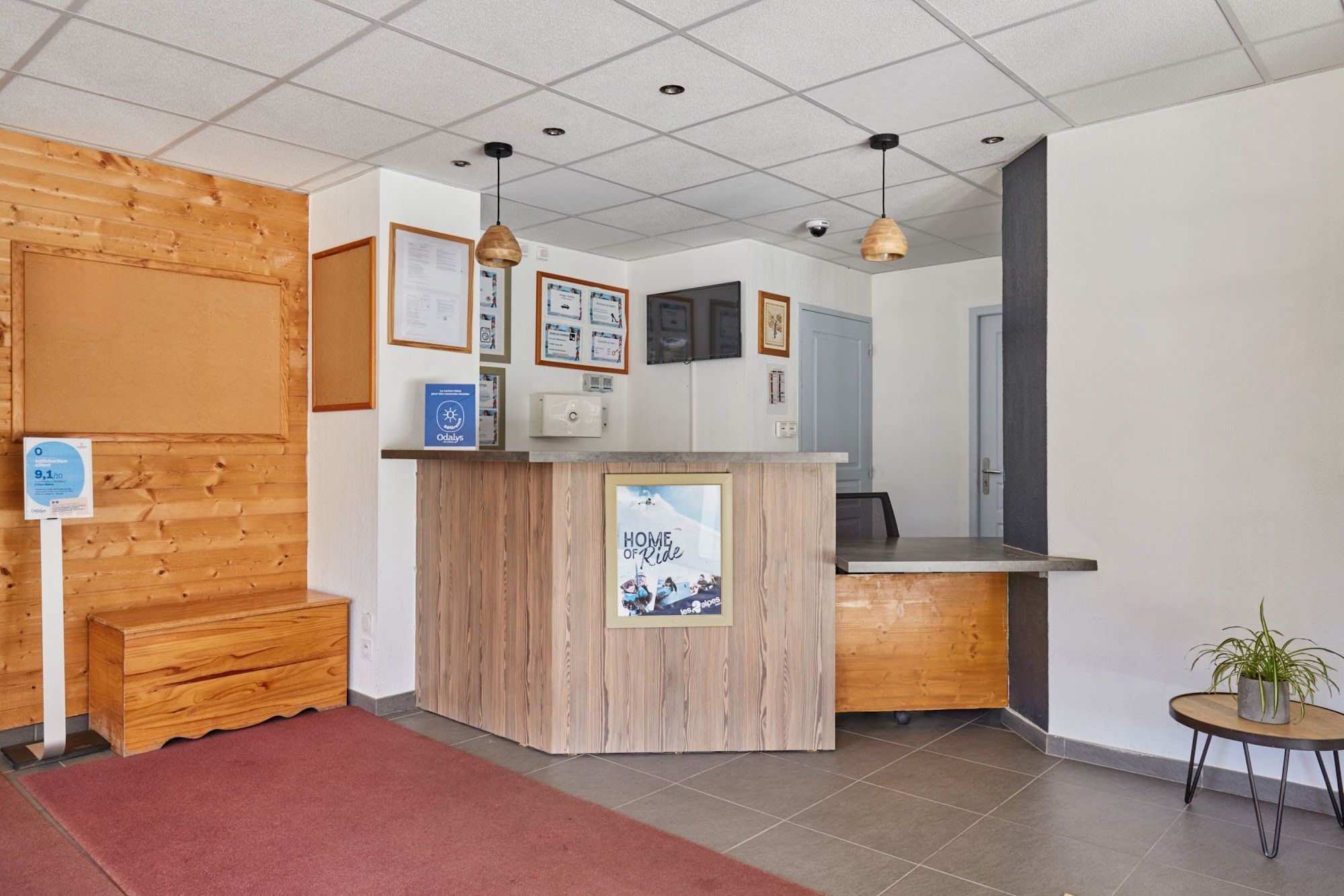 A reception area with a wooden counter, framed pictures, a small table with a plant, and a red carpet.