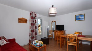 A living room with a red sofa, wooden dining table, chairs, a TV, and a kitchen area with a microwave. - Appart cosy à 150 m des remontées, piscine (Les Deux Alpes)