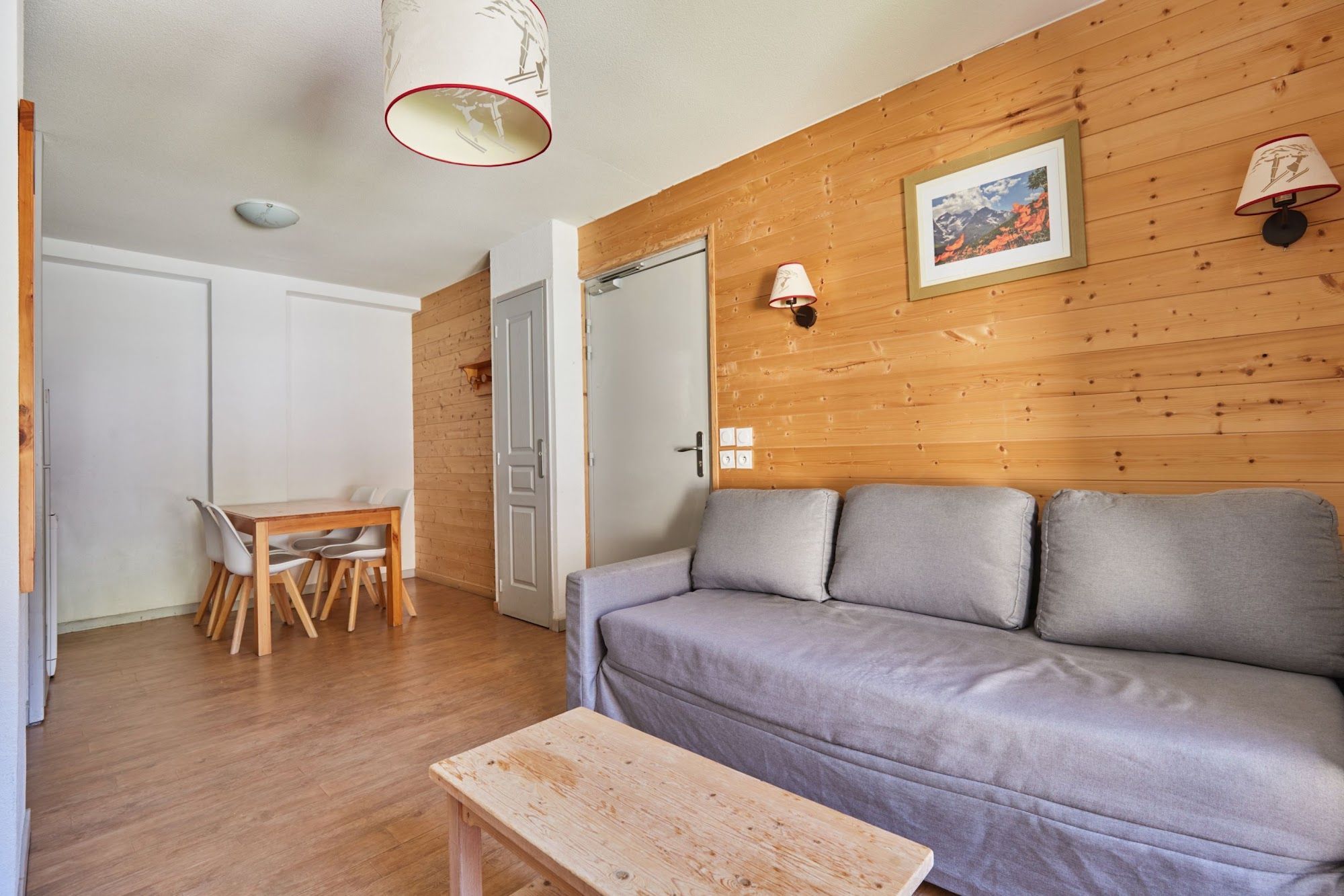 A room with a wooden wall, a grey sofa, a wooden table, and a dining area with chairs.