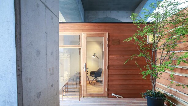 Pared exterior de madera con puerta corrediza de vidrio que da a un espacio interior con escritorio, silla y lámpara.