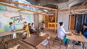 Un café moderno con mesas y sillas de madera, un mural colorido e iluminación que combina luz natural y artificial.