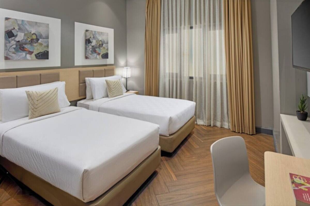 A hotel room with two beds, a desk, a chair, and two abstract paintings on the wall.