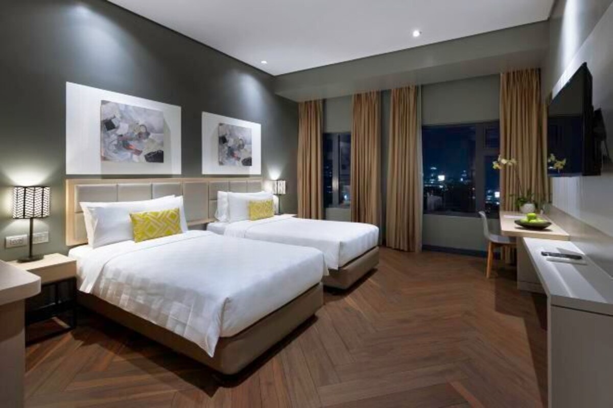 A modern hotel room with two beds, a desk, and a view of the city at night.