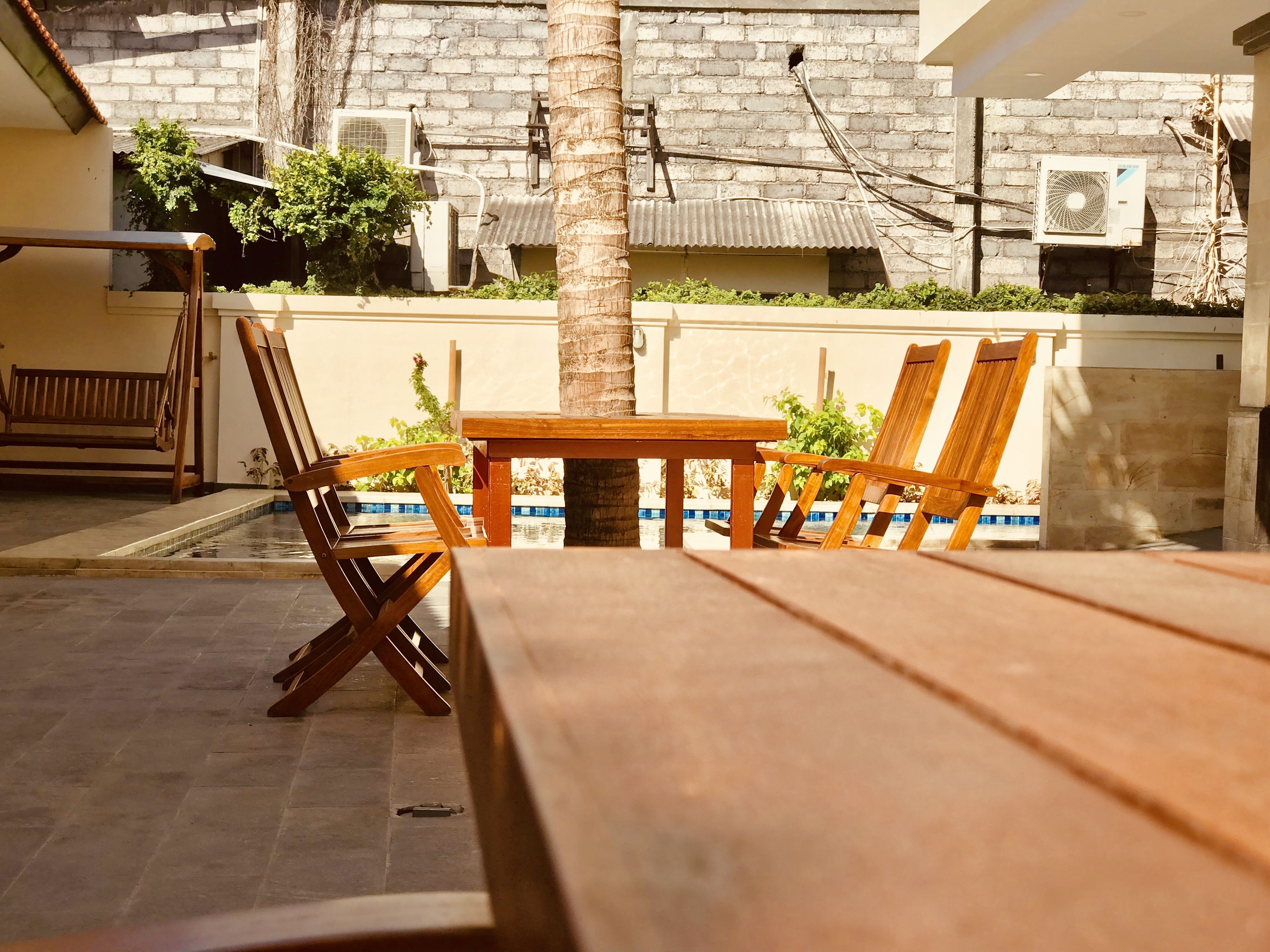 An outdoor patio with a wooden table and chairs, a pool, and a brick wall with air conditioning units.