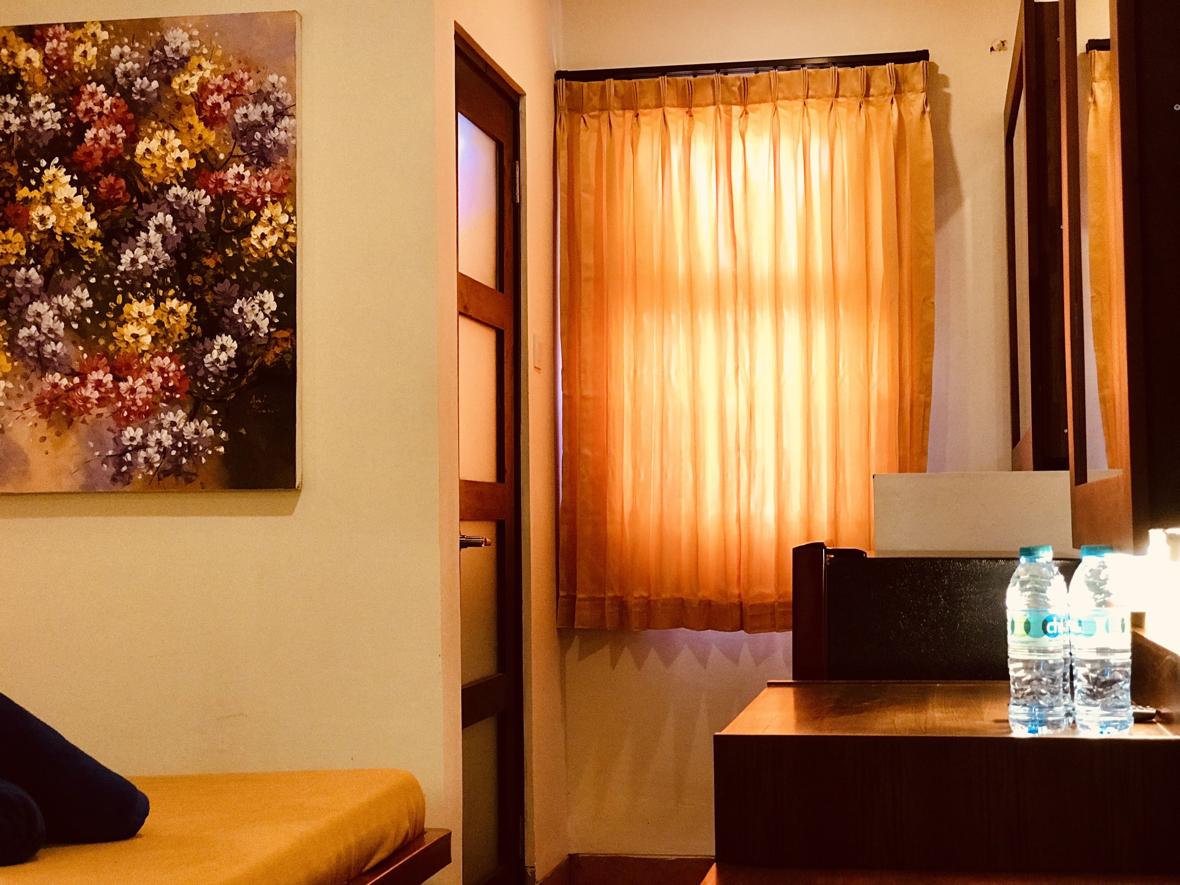 Ein Zimmer mit einem Bett, einem Blumenbild, einem Fenster mit Vorhängen und einem Tisch mit Wasserflaschen.