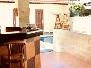 A poolside area with a stone wall, a wooden chair, a table with chairs, and a door in the background. - Familytel Bali (Kedonganan)