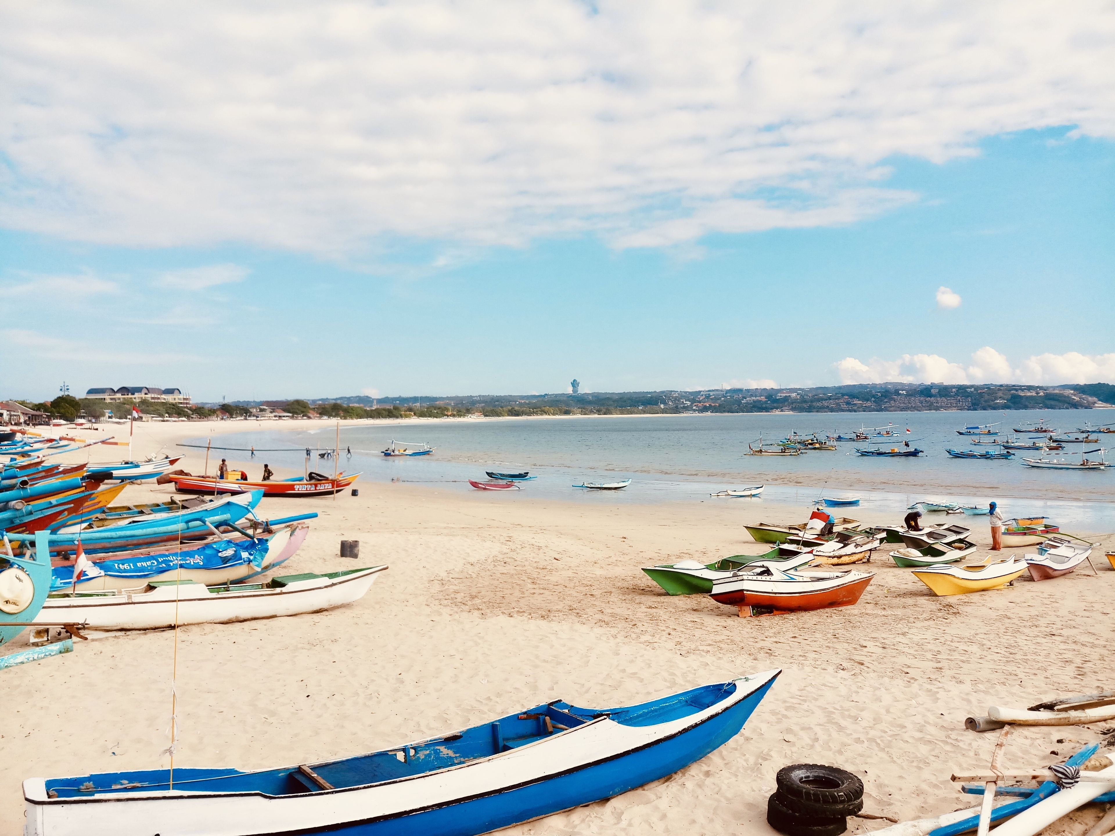 Una playa con numerosos barcos amarrados en la arena y en el agua, cielo despejado y una vista distante de un pueblo.