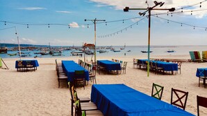 Ein Restaurant direkt am Strand mit Tischen und Stühlen, das einen Blick auf den Strand mit Booten und einem klaren Himmel bietet.