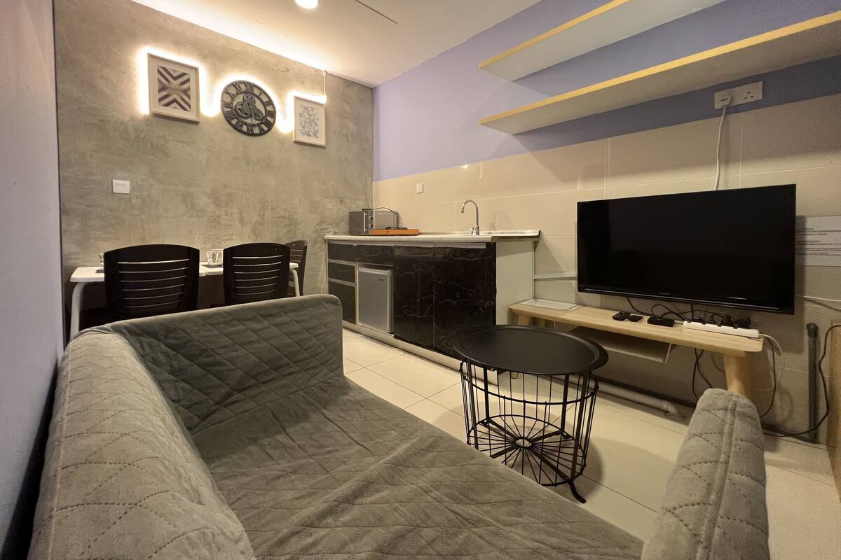 A modern living room with a sofa, a small round table, a flat-screen TV, and a kitchen area with a sink and countertop.