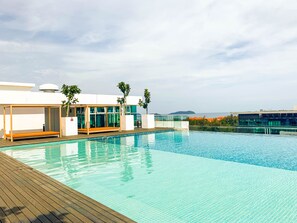 A rooftop infinity pool with wooden deck, glass-enclosed seating, and a view of the sea.