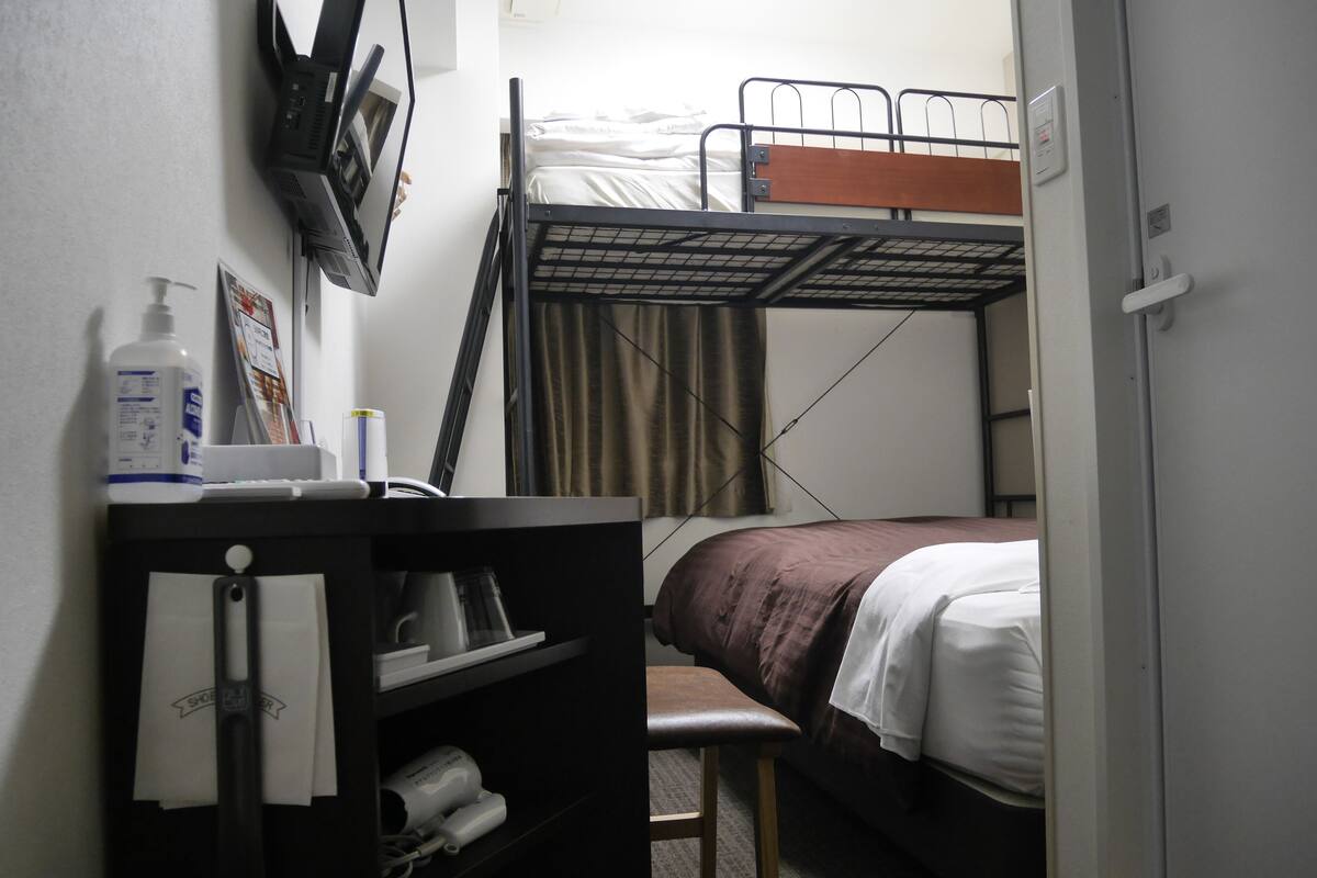 A compact hotel room with a bunk bed, a desk with a chair, a small table, and a shelf with a bottle and a tissue box.