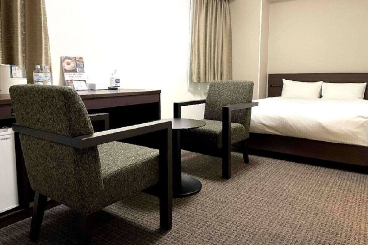 A hotel room with a bed, two armchairs, a small table, a desk, and a window with curtains.