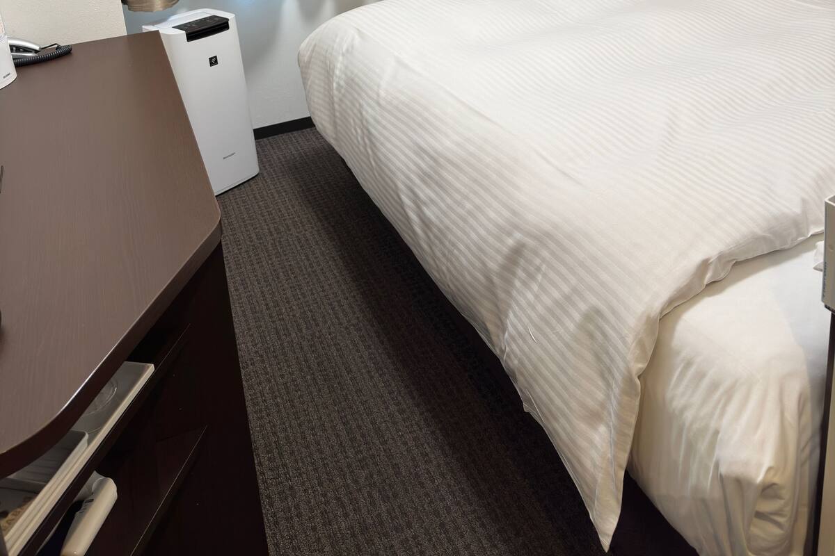 A hotel room with a bed, a flat-screen TV mounted on the wall, a desk with a chair, and a window with curtains.