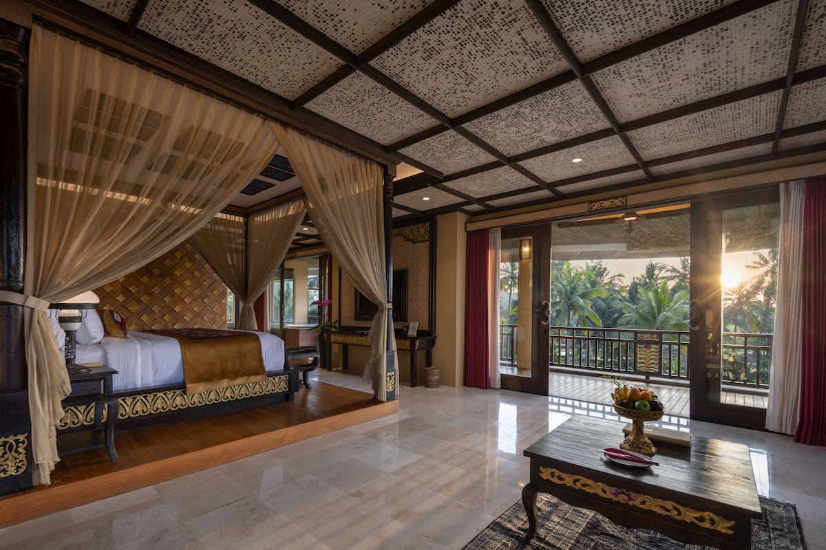 A luxurious bedroom with a canopy bed, a wooden floor, a large window with a view of palm trees, and a balcony with a railing.