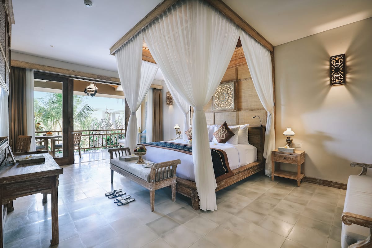 A four-poster canopy bed with a view of palm trees, a wooden desk, and a nightstand with a lamp.
