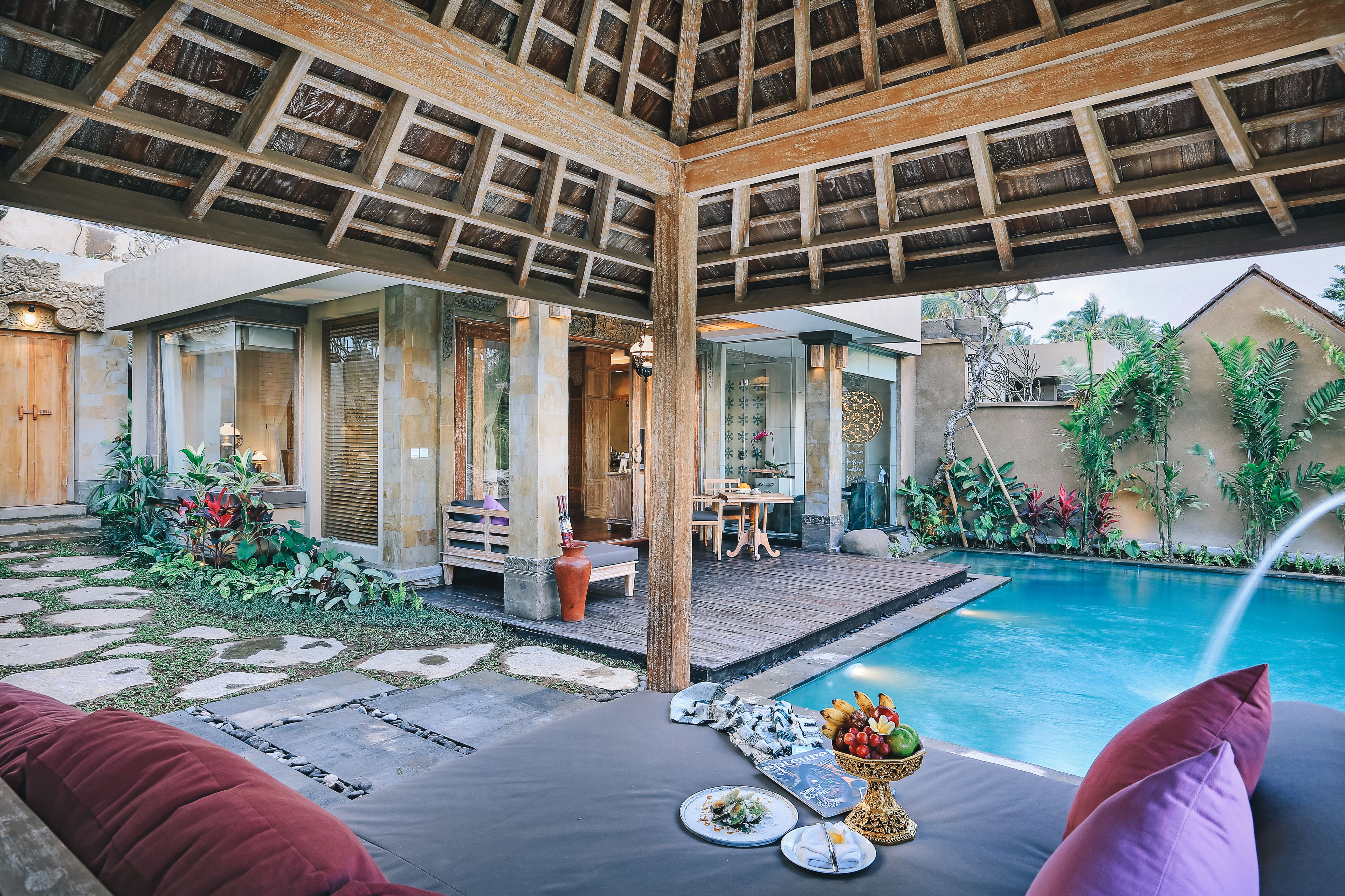 A poolside area with a wooden pergola, stone pathway, and a dining setup with plates and a fruit basket.
