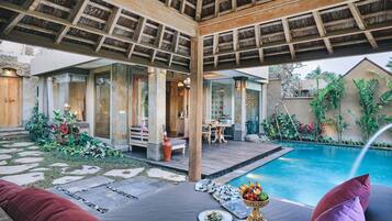A poolside area with a wooden pergola, stone pathway, and a dining setup with plates and a fruit basket.