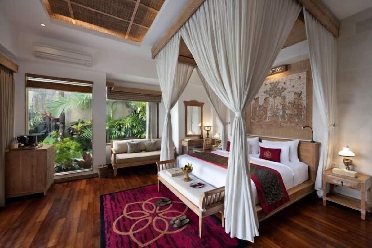 A four-poster bed with a canopy, a wooden nightstand, a bedside table with a lamp, a view of greenery through large windows, and a patterned rug on the floor.