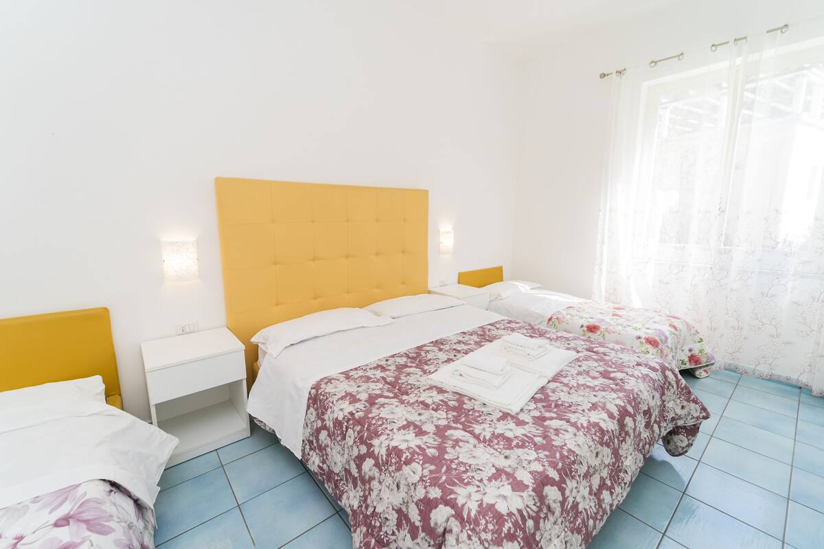 A bedroom with a yellow headboard, two beds with floral bedding, a white nightstand, and a window with sheer curtains.