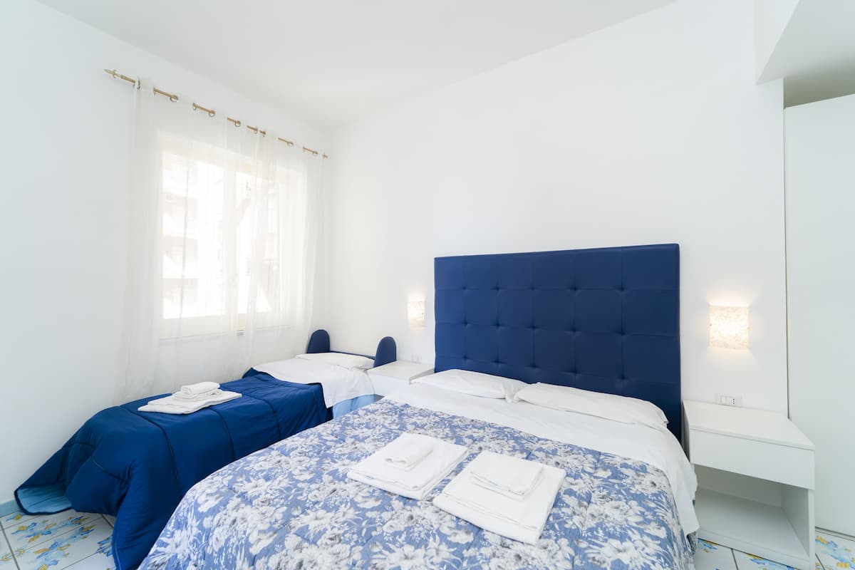 A hotel room with a large bed, two smaller beds, and a window with sheer curtains.