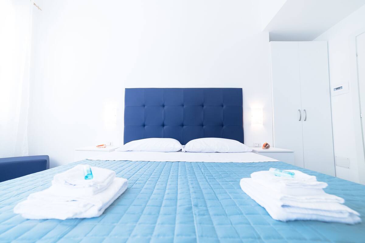 A hotel room with a blue quilted bedspread, white pillows, a tufted headboard, a white nightstand with a phone and a small orange object, and a white wardrobe.