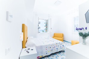 A bright, white room with a bed, a yellow chair, a small table, and a window with a view of buildings. - La Casa di Aurora (Maiori)