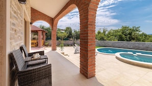 Une terrasse couverte avec des meubles en osier, une colonne en briques et une piscine.