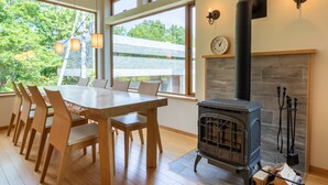 Un comedor con mesa y sillas de madera, un estufa de leña y grandes ventanas con vistas al verdor.