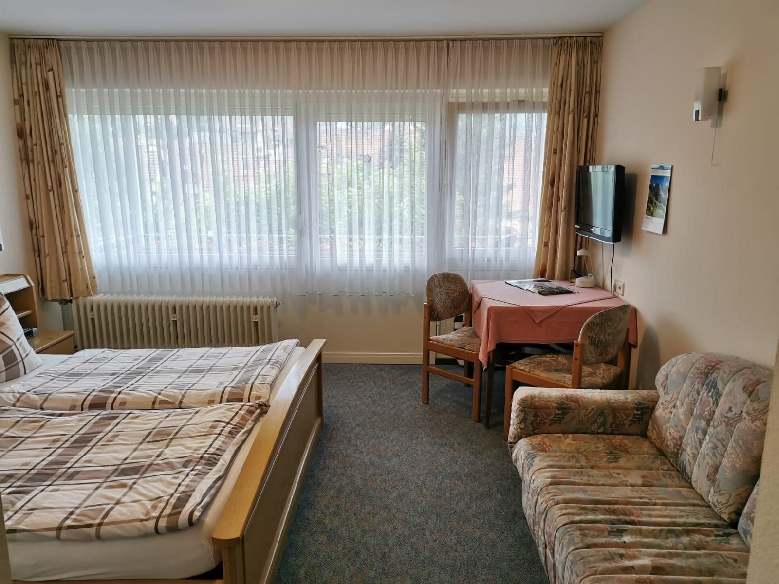 Une chambre d’hôtel avec deux lits, un canapé, une table avec une chaise, un radiateur et un téléviseur fixé au mur.