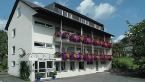 A two-story hotel with balconies adorned with colorful flowers, a sign indicating it is a hotel, and a parking lot in front. - Hartmann (Neuffen)